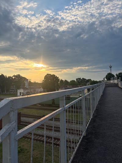 This image shows a view of train tracks at sunset, taken from an elevated position with a metal railing in the foreground. The sun is low in the sky, casting a warm golden light and creating a lens flare. The sky is filled with scattered clouds, some illuminated by the setting sun.

Below the railing, multiple sets of train tracks are visible, laid on gravel beds with some grassy areas between them. A few buildings and trees are scattered along the horizon, suggesting a semi-urban or industrial setting. There is a tall structure resembling a radio tower or industrial chimney in the distance.

The overall mood is serene and tranquil, with the warm light of sunset creating a peaceful atmosphere. The time of day is clearly sunset due to the low angle of the sun and the golden hues in the sky. The location is identified as Romodan, Ukraine. No visible text is present in the image.