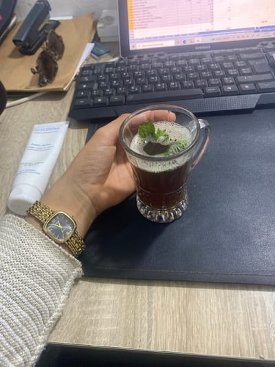 The scene depicts an indoor office or workspace in Monastir, Tunisia, observed from a first-person perspective looking down at a desk. A right hand is centrally positioned, holding a clear glass mug that contains a dark brown liquid topped with a layer of white foam and several green mint leaves. The wrist of the hand is adorned with a gold-toned square-faced watch featuring a black dial, and a light-colored knitted sleeve is visible. The mug rests on a dark blue desk mat. To the left of the hand, a white tube labeled "GLYCERINA" is placed on a light wood-grain desk surface. In the background, a black computer keyboard with Arabic characters is visible, positioned in front of a silver Samsung laptop displaying a spreadsheet application with data and text in Arabic script. Further to the upper left, a pair of sunglasses lies on a piece of brown paper next to a black stapler.