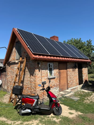 The image depicts a red electric scooter parked on a dirt and grass area in front of a single-story brick building. The building has a sloped roof covered with solar panels, a brown metal chimney, and two brown metal doors. A wooden ladder is propped against the left side of the building. A black storage box is attached to the rear of the scooter. The scene appears to be a residential setting in the village of Zmiinets, Ukraine. The sky is clear and blue.