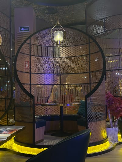 The image shows a dining booth in a restaurant. The booth is circular in shape and made of decorative metal screening. Inside the booth, there is a table and seating upholstered in dark material. A pendant light hangs above the table. The booth is illuminated by yellow lighting around its base and also by ambient colored lighting from within the restaurant. In the foreground, a dark chair is partially visible. A sign with a white arrow pointing left on a green background is mounted on a pillar to the left of the booth. The overall aesthetic suggests an Asian-inspired decor. The restaurant is located on Carretera De San Martín, Spain.