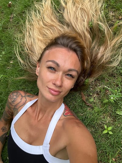 A woman is lying on the grass, facing the camera. She has long, blonde hair spread out around her head, and her eyes are open and looking at the camera. She is wearing a black and white tank top. Tattoos are visible on her arm and shoulder. The grass is green and lush, with some fallen leaves scattered around. The scene appears to be outdoors, possibly in a park or backyard, in Thôn 2, Vietnam.