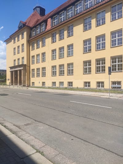 This image shows a large, yellow, multi-story building with a red-tiled roof and a dark grey turret, located on the street in Chemnitz, Germany. The building has numerous windows with white frames. In the foreground, there is a paved road with white lane markings and a sidewalk. A blue parking sign with a car symbol is visible on the right side of the building. The sky is blue with some white clouds.