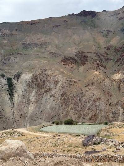 A serene landscape unfolds in Reru, India, showcasing a tranquil pond nestled at the base of a vast, arid mountain. The mountain, a dominant feature, displays a rugged texture of brown and gray rock, interspersed with patches of sparse green vegetation clinging to its slopes. The sky above is overcast, casting a soft, diffused light over the scene.

In the foreground, a winding dirt road curves gently towards the pond, bordered by dry, yellowish grass and scattered stones. A large, weathered boulder rests on the left, its rough surface weathered by time. A low stone wall, constructed from smaller rocks, forms a boundary on the right side of the road, with another large, dark-colored boulder prominently placed behind it.

The pond itself is a body of still, light-green water, reflecting the muted tones of the sky and surrounding terrain. A single, slender utility pole stands near the edge of the pond, its dark silhouette contrasting with the pale water. Two small, dark-green trees are visible on the far bank of the pond, adding a touch of life to the otherwise austere environment. The overall impression is one of natural beauty and quietude, a testament to the stark yet captivating landscapes of Reru.