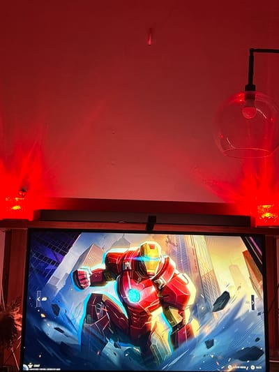 The image shows a wall illuminated by red light, with a glass orb lamp attached to the wall on the left. A television displaying an image of Iron Man in a dynamic pose with city buildings in the background is positioned on the right. The lower portion of the screen displays some text. Small, square, illuminated objects are positioned vertically along the edge of the television frame. The scene appears to be indoors.