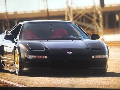 This is a forward-facing, eye-level shot of a black Honda NSX sports car. The car is parked on an asphalt road with a slight curb in the foreground. The background consists of a blurred industrial structure, possibly a bridge or overpass, under a bright, possibly daytime sky. The car's interior, visible through the windshield, features two red racing seats. The car has gold rims on its wheels. There is a red and white Honda emblem on the front of the car. No people are visible. The overall mood is one of power and sleekness.