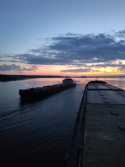 The image captures a twilight scene on a body of water, likely a river, near Irdomatka, Russia. A barge, labeled "КУНЦЕВО" on its side, travels along the water. Another larger barge is closer to the camera, taking up the right side of the frame, blocking the view. The sky is a mix of blue, orange, and grey, indicating a sunset or sunrise. The water reflects the colors of the sky, and the surrounding land is dark and indistinct.