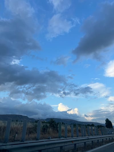 The image depicts a landscape scene with a cloudy sky over rolling hills. In the foreground, there is a guardrail and a series of concrete posts bordering a dry, grassy area. Behind this, a fence is visible, with sparse vegetation and some trees. The hills in the background are covered in a muted green and brown landscape, with some structures or buildings interspersed. The sky is a mix of blue and various shades of gray clouds, with some patches of bright white clouds suggesting sunlight breaking through. The overall lighting indicates it is likely late afternoon or early evening. There are no people or vehicles visible in the scene. The location is identified as Manfredonia, Italy.