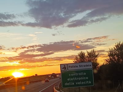 The image captures a dramatic sunset over an Italian highway, likely near Somaglia, Italy. The sky is ablaze with vibrant oranges, yellows, and pinks, with dramatic clouds casting long shadows and reflecting the intense light of the setting sun. The sun itself is a brilliant orb, partially obscured by the horizon, casting a warm glow across the scene.
In the foreground, a green road sign with white lettering indicates "Polizia Stradale" (State Police) at the top, with a symbol of a police officer. Below this, in larger white text on the green sign, it reads "controllo elettronico della velocità," which translates to "electronic speed control" or speed camera. This suggests the presence of speed enforcement measures on this stretch of highway.
The highway itself is visible, with multiple lanes and guardrails. Cars are seen driving on the road, their headlights on in the fading light. The setting is rural, with trees lining the side of the road, silhouetted against the colorful sky. The overall mood is serene yet dynamic, with the beauty of the sunset juxtaposed with the functional signage of the highway. The time of day is clearly sunset, and the weather appears clear, allowing for the spectacular display of colors in the sky.