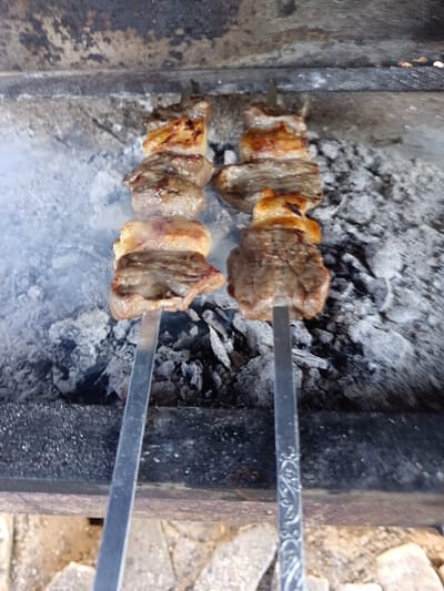 The image displays two metal skewers, each laden with alternating pieces of browned meat and golden-brown rendered fat, positioned horizontally over a bed of glowing charcoal embers and white ash within a stone or metal brazier. Wisps of white smoke rise predominantly from the left side of the embers, indicating active grilling. The visible end of the right skewer features an etched decorative pattern. The immediate foreground reveals the dark, textured rim of the brazier, resting on an irregular stone or paved surface. This scene is situated in Kasri Arifan, Uzbekistan.
