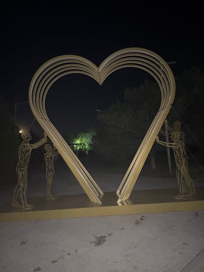 In Egorlykskaya, Russia, a large, golden, heart-shaped sculpture is illuminated at night. The sculpture is composed of multiple concentric heart outlines. Two wireframe figures, one taller on the left and one shorter in the center, appear to be supporting the left side of the heart. On the right side, a similar wireframe figure is depicted supporting the right side of the heart. The figures are stylized and appear to be made of thin metal rods. The sculpture is situated on a paved area, possibly a walkway or plaza, with sparse vegetation growing in the cracks. In the background, trees are visible, and a faint green light emanates from an unseen source, possibly a spotlight on the trees. Streetlights are also present, casting light on the scene. The overall impression is of a public art installation at night.