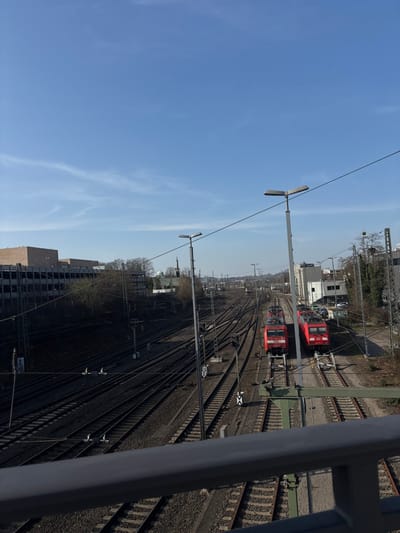 The image depicts a wide expanse of railway infrastructure in Aachen, Germany, viewed from an elevated perspective, likely a bridge, with a dark grey handrail partially obscuring the lower foreground. The scene is illuminated by bright daylight under a clear, predominantly blue sky with sparse, wispy cirrus clouds.
In the middle ground and extending into the background, a complex network of numerous parallel railway tracks, switches, and sidings stretches towards the horizon. The tracks are laid on ballast. Overhead catenary lines and supporting poles are visible throughout the railway corridor. Several grey street light poles with 'V'-shaped twin luminaires are prominent, particularly in the mid-ground. Black and white signal markers are positioned between some tracks.
Two red electric locomotives, identified as DB Class 185 series, are positioned stationary on separate tracks to the right of the frame, facing towards the distant horizon. The locomotive on the left is partially obscured by a streetlight pole. No people are visible.
To the left of the railway tracks, a multi-story building with a dark facade and lighter upper section (possibly a parking garage or industrial complex) is visible behind a line of bare trees and dense vegetation. Further in the mid-ground left, a distinct white building with a green domed roof, identifiable as the Greek Orthodox Church Agia Ekaterini, is visible above the tree line. To the right of the tracks, several industrial or commercial buildings with flat roofs are present, along with a partially visible parking area containing several vehicles. The surrounding vegetation consists of bare trees and shrubs, suggesting a season outside of full foliage. The railway lines converge into the distance under a slightly hazy horizon.