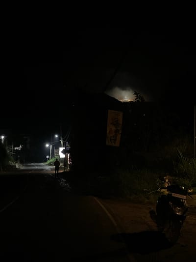 The image shows a night scene in Kumbalwela, Sri Lanka. A person is walking on the right side of a road, lit by streetlights. A motorcycle is parked on the side of the road near foliage. A sign is visible in the distance. Dark buildings border the left side of the road.