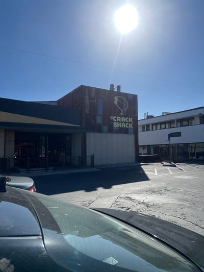 The image shows the building of "Crack Shack", a restaurant known for its chicken dishes, in Costa Mesa, California. The building has a weathered, rusty appearance and the restaurant's logo prominently displayed on it. A car is partially visible in the foreground, with the restaurant situated on the left side of the frame. The parking lot and an adjacent building are also in view. The bright sunlight illuminates the scene.