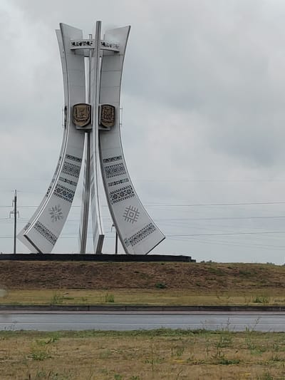 An outdoor, eye-level, full shot shows a large, white, modern monument located in a grassy area with a road in the foreground. The monument is tall and has a symmetrical design, with four curved vertical elements that widen at the bottom. Each of the four elements is decorated with intricate, geometric patterns in black and blue, reminiscent of traditional embroidery. Two prominent, shield-shaped emblems in a metallic bronze color are mounted on the front of the monument, one on each of the two central vertical sections. Above these emblems, the monument splits into four thinner vertical sections that form a cross-like structure at the very top, with a circular platform visible just below the apex. The monument stands on a raised, brown earthen mound, with a clear sky with light clouds overhead. Power lines are visible in the background, stretching across the sky. The foreground features a paved road with grassy verges on either side, suggesting the monument is a roadside landmark. The overall impression is one of a significant civic monument situated in an open, public space, likely within or near the city of Brest, in the village council of Charnaučycki selski Saveet, Belarus.