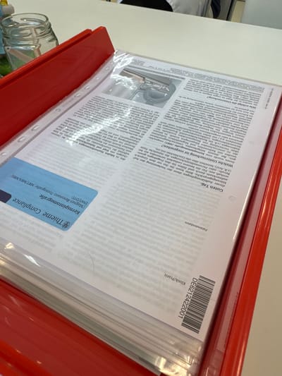 The image shows a red binder containing documents related to a magnetic resonance imaging (MRI) scan. The documents include patient data, a clinic/practice identifier, and information about the MRI procedure, including instructions and potential risks. A blue label with "Thieme Compliance" and "Kernspintomografie" is visible. The barcode on the document is "DE6212422001". A glass jar with a green lid is partially visible in the lower left corner of the frame. The document is in German. The city of Amstetten, Austria is not identifiable from the contents of the image.