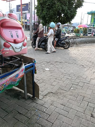 The scene is an outdoor paved area in the city of Sumberejo, Indonesia. In the immediate left foreground, a pink and white children's ride resembling a cartoon train or vehicle with a smiling face is present, mounted on a blue metal frame with a colorful banner. The ground is paved with grey interlocking bricks laid in a herringbone pattern, showing scattered debris.

In the midground, three individuals are situated with two dark-colored scooters. A person wearing a brown long-sleeved shirt, beige hijab, light pants, and sandals is seated on the left scooter, facing right, with hands on the handlebars. To their right, a second person wearing a light-colored checkered top, light pants, and a light blue helmet stands beside a second scooter, holding a smartphone and facing left towards the first person. A third person, wearing a dark jacket, shorts, and a white helmet, is seated on the second scooter behind the standing individual, also facing right.

The background features a large green-leafed tree. Beyond the tree, a concrete planter box or curb separates the paved area from a dirt patch. Further back, a green and white structure, possibly a building or shelter, is visible on the far right, with a colorful striped umbrella adjacent to it. In the distant background on the left, utility poles and lines are present, along with a sign displaying "Mandiri ATM."