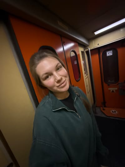 A young woman smiles slightly, wearing a dark green zip-up jacket over a black top. She appears to be inside a train car with orange walls, recognizable by doors labeled with numbers. Artificial light illuminates the train's interior. The photograph is angled, giving a slightly skewed perspective of the scene.