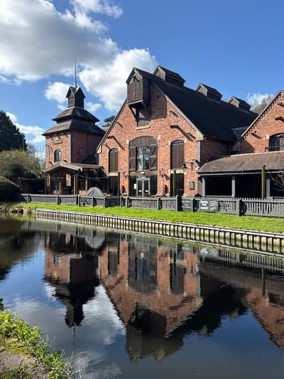 The scene depicts The Watermill, identified as the Old Fulling Mill, situated alongside the Staffordshire and Worcestershire Canal in Stourport-on-Severn, within the Wyre Forest district of the United Kingdom.
The primary subject is a multi-section structure constructed from reddish-brown brick with dark-tiled roofs. The left section features a square, two-tiered tower with a pointed spire. The central and right sections consist of gabled buildings with large dark-framed windows, some featuring wooden shutters, and several wooden dormer windows protruding from the roofline. A covered outdoor patio, supported by wooden posts, extends from the right side of the main building. A dark grey picket fence lines the edge of the property facing the canal, separating it from a narrow strip of green grass. A black A-frame sign reading "WELCOME TO THE WATERMILL" is positioned behind the fence. No individuals are visible.
In the foreground, a calm body of water, reflecting the entire building complex, the fence, and the sky, occupies the lower half of the image. The water's surface exhibits a ripple pattern from the reflections. The sky overhead is bright blue with scattered white cumulus clouds. Deciduous trees and green foliage are visible on the left and in the background, suggesting a natural environment surrounding the establishment. The scene is illuminated by strong sunlight, creating clear shadows.