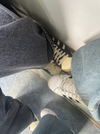 The image shows a close-up, overhead view of four legs wearing denim jeans and athletic shoes. Two pairs of legs are visible in the foreground, one on the left wearing dark wash jeans and black and white Adidas sneakers, and another on the right wearing light wash jeans and white Adidas sneakers. Behind these, portions of two more legs wearing dark wash jeans and white athletic shoes are partially visible. The individuals appear to be sitting. The environment is utilitarian, with a grey, textured floor and a white wall or surface in the background, suggesting an indoor, possibly public space. The image does not provide any specific visual cues to definitively place the scene in Aprelevka, Russia; its context is a generic urban or indoor setting.