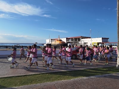 A group of twenty-five people, primarily young adults, are walking in a line down a sidewalk near the ferry terminal in Cozumel, Mexico, located at coordinates 20.511, -86.95. Most of the people are wearing pink shirts and white shorts, and some are carrying drums. The sidewalk is made of concrete and brick, with a patch of green grass growing along its edge. The group is walking towards a large orange ferry boat, which is docked behind them. A red car is parked on the sidewalk to the right of the group. The sky is blue and sunny. The air appears clear and bright. The environment is hot and humid.