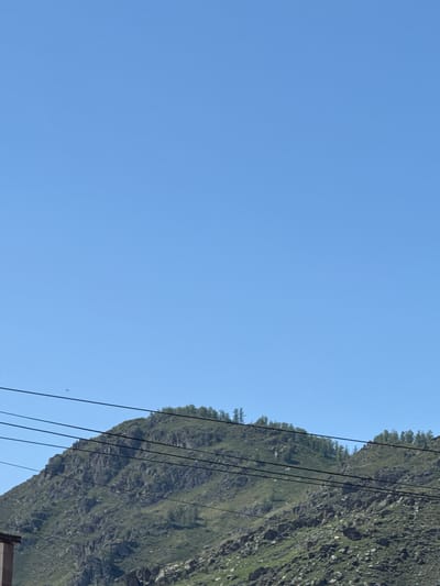An outdoor, vertical, medium shot captures a clear blue sky above a tree-covered, rocky mountain in Onguday, Russia. The mountain occupies the bottom half of the frame, its slopes covered in a mix of green grass and large, gray rocks. A line of trees runs along the crest of the mountain. Several black power lines stretch horizontally across the lower portion of the image, intersecting the mountain and sky. A small, dark object, possibly a bird or drone, is visible in the sky to the left of the mountain. In the lower left corner, a small section of a brown building is visible. The scene is well-lit, suggesting daylight.