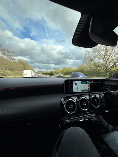 The view from inside a car shows a highway in Mid Suffolk, United Kingdom. The car's dashboard is visible, including air vents and a navigation screen displaying a map. Outside, traffic is present, with vans and cars on the road. The sky is partly cloudy. A contrail is visible. Trees line the highway. The driver is wearing denim jeans and athletic shoes.