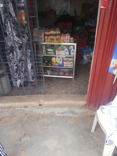 This image depicts the exterior of a small roadside shop in Ajasse Ipo, Nigeria. The shop appears to be a kiosk or a stall with a red metal door on the right. Inside, shelves are stocked with various goods, including what appear to be bread loaves, packaged snacks, and beverages in cartons and bottles. There are also crates of drinks visible behind the shelves. A person is partially visible behind a metal grille in front of the shop, wearing a patterned fabric. The ground in front of the shop is a rough, concrete surface. A white plastic chair with some fabric draped over it is positioned to the right of the shop entrance. The overall scene suggests a typical small business operation in a Nigerian town.