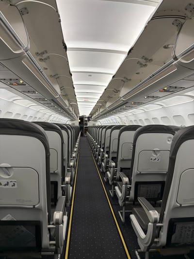 This is an interior shot of an airplane cabin, most likely taken during boarding or deplaning, as no passengers are visible. Rows of seats line both sides of a central aisle carpeted with a dark-patterned design, featuring a yellow stripe on either side. The seats are predominantly gray with dark headrests, and each has a tray table in the upright position. Overhead compartments run along the length of the cabin, with reading lights and air vents visible above each row of seats. The overall lighting is bright, creating a clean and sterile environment. The image is taken from a slightly elevated perspective, looking down the length of the airplane. There is nothing indicating the image was taken in Communauté rurale de Diass, Senegal.