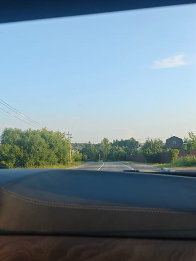 The image is a first-person perspective from inside a vehicle, looking forward through the windshield. The foreground is dominated by the dark, textured dashboard of the car. Beyond the dashboard, a two-lane asphalt road stretches into the distance, curving slightly to the right. The road has white lane markings.

To the left of the road, there is a dense cluster of green trees and foliage, with a utility pole and power lines running parallel to the road. Further into the distance, more trees and vegetation line the sides of the road, with some houses visible through the greenery. One house, with a dark brown, sloped roof and a single window, is prominent on the right side, partially obscured by a wooden fence and climbing plants.

The sky is a clear, light blue, with a few wispy white clouds scattered across it. The overall scene depicts a rural or suburban landscape in daylight, likely during the summer given the lush greenery. The location is identified as Венюково Снт, Russia.