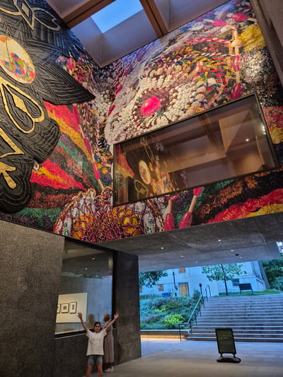 In Princeton, United States of America, a young girl with arms outstretched stands in a modern architectural space with a large, colorful mosaic mural adorning the walls. The mural features intricate patterns and vibrant colors, with a prominent pink circular element. Above the mural, a skylight illuminates the area. To the right of the mural, a large glass-encased opening reveals more of the artwork. In the foreground, a woman in a light-colored dress stands behind the girl. The space appears to be an atrium or entranceway, with granite-like textured walls and a concrete ceiling. Beyond the entryway, a set of outdoor stairs leads up to a building surrounded by greenery and trees. A sign at the bottom of the stairs advertises a "Summer Celebration" event. The overall scene captures a blend of art, architecture, and a lively outdoor setting.