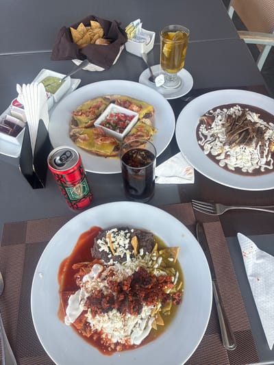 The image shows a tabletop scene with various dishes and beverages, likely in Pueblo Nuevo de Jasso, Mexico. Three plates of food are visible: one with enchiladas covered in mole sauce and sprinkled with cheese and onions, another with enchiladas possibly filled with meat or cheese and topped with a small container of pico de gallo, and a third with chilaquiles (tortilla chips simmered in salsa) accompanied by beans, chorizo, and sour cream or cheese. A basket of tortilla chips with containers of guacamole is present. Beverage items include a glass of iced tea, a glass of cola, and a can of soft drink. Cutlery, napkins, sugar packets, and condiments are arranged around the dishes. The table surface is dark gray, with a brown textured placemat on the right. The overall setting suggests a meal at a restaurant or outdoor dining area.