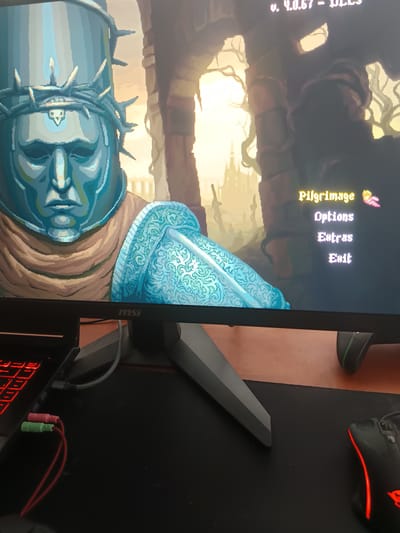 The image shows a close-up view of a computer monitor displaying the main menu of a pixel-art video game. The game's title, "Pilgrimage," is prominently displayed in yellow, followed by menu options like "Options," "Extras," and "Exit." The background art depicts a character with a large, ornate helmet adorned with thorns, and the overall aesthetic is dark and medieval. The monitor is set on a desk, with a laptop and gaming peripherals visible in the foreground, including a mouse and audio jacks. There is no indication of Lyubimets, Bulgaria, or any specific city in the image.