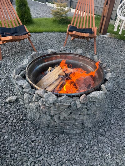 This image depicts an outdoor scene at a location in Sharovechka, Ukraine, featuring a fire pit surrounded by dark gray gravel. The fire pit is constructed with a metal basin filled with burning wood and logs, emitting bright orange flames and smoke. The exterior of the fire pit is a gabion cage filled with gray stones. In the background, two wooden chairs with black cushions are positioned on the gravel. A small evergreen tree and a patch of green grass are also visible, along with a fence in the distance. The overall environment suggests a backyard or patio setting.