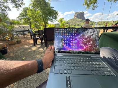 A first-person perspective shot shows a person using a Lenovo Yoga 7i laptop outdoors. The screen displays a desktop with numerous icons and a background of trees with a purple and pink sky. The person's left arm, adorned with a black smartwatch, is visible in the foreground.
In the background, a verdant landscape is visible, suggesting a rural or natural setting. A large rock formation, likely Sigiriya Rock, is present in the distance. Other people can be seen seated at a table in the background, engaged in conversation and activities. The outdoor seating area features wooden tables and chairs, with potted plants adding to the ambiance. The overall atmosphere suggests a relaxed setting in Sigiriya, Sri Lanka.