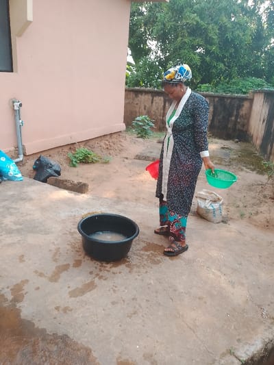 This image captures an outdoor domestic scene in Ufuma, Nigeria. A woman, appearing to be pregnant, stands with her head bowed, looking down. She wears a dark floral print robe tied at the waist over green and blue patterned trousers, and black double-strap sandals. A multi-colored headwrap with partial text "UNDER" is on her head. She holds a green plastic colander in her right hand. On the concrete ground in the foreground, a black plastic basin contains a cloudy liquid. Water splashes and wet marks are visible on the concrete around the basin. To the woman's left, a red plastic bowl rests on sandy ground. To her right, a white sack, possibly containing cement, is visible on the sand. The background features a light peach-colored building wall on the left, equipped with a white PVC pipe system. A blue plastic bag and a black plastic bag are positioned near the base of this wall. A low brown wall runs across the mid-ground. Green foliage, including small plants and a large tree, is visible behind the walls and in the sandy areas.
