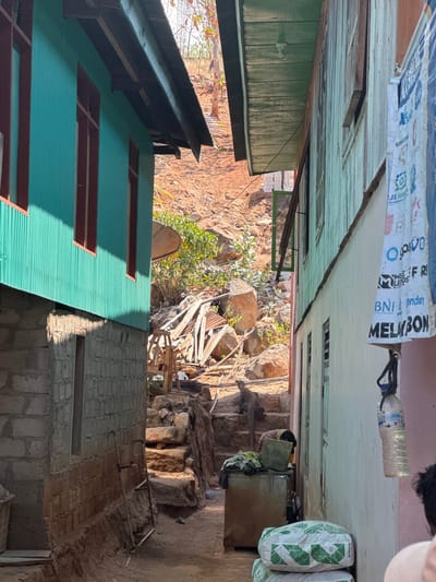 This is a close-up, eye-level, vertical shot of a narrow, unpaved alleyway between two residential buildings in Komodo Village, Indonesia. The building on the left has turquoise corrugated metal siding and red-trimmed windows, with a grey concrete block foundation. The building on the right has weathered, light green wooden siding. The ground is a mixture of dirt and rough stone steps leading up a hillside in the background, which is a reddish-brown earth slope covered with rocks and sparse vegetation.
In the foreground, on the right side of the alley, there are two large, white bags with green geometric patterns, likely containing cement, stacked on the ground. A plastic water bottle hangs from a string attached to the right building. Further back, there is a rusted metal cabinet with clothing and a green bag on top. Several wooden planks and corrugated metal sheets are piled haphazardly on the right side of the alleyway, suggesting construction or disrepair.
A medium-sized, grey lizard is perched on the stone steps in the middle ground, facing away from the camera. A person's head and shoulder, wearing a light-colored shirt, are visible in the lower right corner of the frame, facing into the alleyway. The lighting suggests it is daytime, with strong shadows cast by the buildings and objects. The overall scene depicts a humble, possibly developing, residential area with signs of everyday life and rudimentary construction.