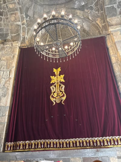 A dark burgundy curtain adorned with a gold, ornate cross sits against a stone wall. The curtain features a decorative border along the right edge. To the left, a complex iron chandelier with numerous lights illuminates the scene. The room appears to be a church or similar religious space, with visible stone arches and walls.