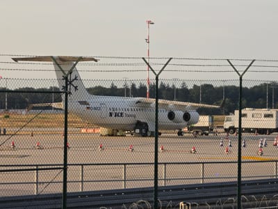 The image shows an airport tarmac in Kelsterbach, Germany, with a white airplane labeled "D-NICE ICE Training" as the primary subject. The airplane has two engines under its wings and a distinctive snowflake logo on its tail. It is parked on the tarmac, surrounded by various airport vehicles, including a white truck and a mobile command center. Traffic cones and lights are placed around the tarmac, indicating an active airport environment. In the background, a line of trees forms a backdrop, and a red and white striped pole with lights and a weather sensor stands tall. The scene is captured through a chain-link fence with barbed wire on top, suggesting a restricted area. The sky is overcast, and the lighting suggests it is either early morning or late afternoon. There are no people visible in the image.