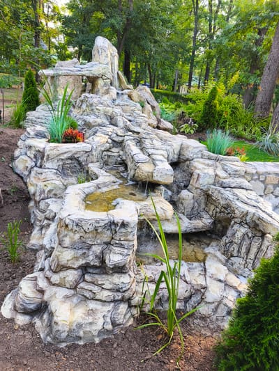 The image shows a man-made rock waterfall feature in a garden setting. The waterfall is constructed from large, irregularly shaped rocks that are layered to create multiple tiers and cascades. Water flows down the rocks into shallow pools at different levels. Green plants, including ornamental grasses and a small coniferous shrub, are planted around the base of the waterfall and in small crevices on the rocks. In the background, there is a dense line of mature trees. The ground around the waterfall is bare earth with some fallen leaves. The scene appears to be in a park or landscaped garden. The location is identified as Stroiesti, Moldova. There are no people visible in the image.