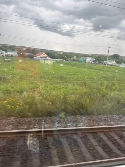 The image depicts a rural landscape with a village and train tracks. The sky is overcast with heavy gray clouds. In the foreground, train tracks with ballast are visible. Beyond the tracks, a grassy field with scattered wildflowers stretches towards a cluster of houses. The houses have various colored roofs, including red, green, and white. Trees and bushes surround the village. In the distance, a gentle hill is visible. The overall scene suggests a cloudy day in a rural settlement, likely in Russia, specifically Kurkachi.