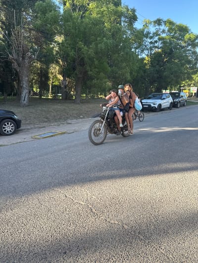 Several people are riding a motorcycle on an asphalt road. Trees and parked cars are visible on the side of the road. The day is sunny.