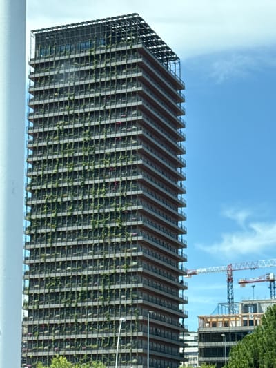 The media file depicts a tall, modern building with a facade covered in greenery. The building's design features a stepped or tiered structure, with horizontal elements that create a distinct visual pattern. The greenery appears to be planted in a way that cascades down the sides of the building, giving it a vertical garden aesthetic.
In the background, construction cranes are visible against a bright blue sky with scattered clouds, suggesting ongoing development or renovation in the area. The presence of cranes and unfinished structures indicates a construction site.
The overall scene appears to be an outdoor urban environment, likely during the daytime given the bright sky and clear visibility. The weather seems pleasant, with a clear sky suggesting good conditions. The location is identified as Esplugues de Llobregat, Spain. No people are clearly visible in the frame. The building itself, with its innovative design incorporating nature, suggests a focus on sustainability or a contemporary architectural style.