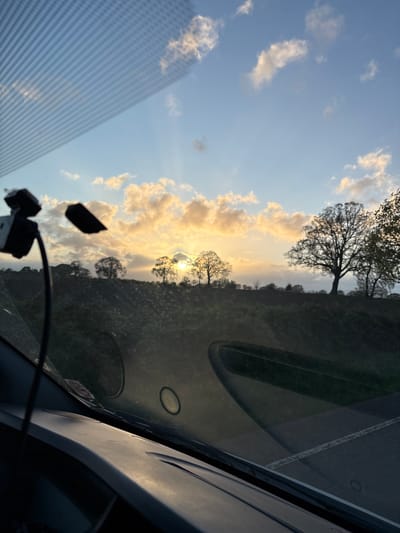The image, taken from inside a vehicle, shows a view through the windscreen of a rural landscape under a partly cloudy sky with the sun low on the horizon. The location is likely Ruyton-XI-Towns, United Kingdom, based on user information. Several trees are silhouetted against the sun, indicating the approximate location of a treeline or forest. The interior of the car, including the dashboard, speakers, and some mounted electronic equipment, is visible. The light suggests it is either sunrise or sunset. No people are directly visible in the frame.