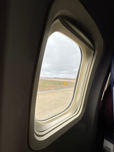 The image displays a view from inside an aircraft, looking out through a passenger window at an airport in Nashville, United States of America. The window features a light beige frame with a multi-layered appearance and a metallic element along its top edge, possibly part of a shade mechanism. To the immediate left, a dark grey interior panel of the aircraft is visible. To the right, a portion of a passenger seat back, appearing dark blue and maroon, is present. Outside the window, a concrete airport tarmac with parallel double yellow lines is visible in the foreground. Beyond the tarmac, a strip of dry, light green and brown grass extends. In the midground, a yellow square sign displaying "S6" in black text is positioned on the grassy area. The sky above is overcast and uniformly grey. The aircraft appears to be stationary on the ground. No individuals are visible within the frame.