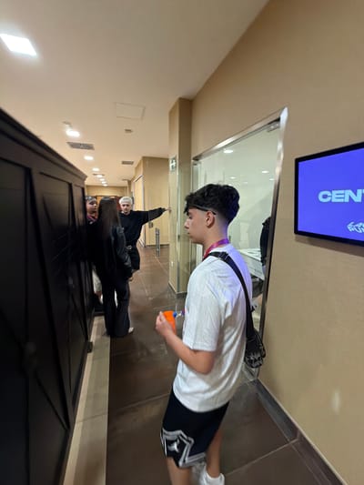 A hallway scene shows a young man in a white patterned shirt, black shorts with a white logo, and white sneakers. He wears glasses and has a red lanyard around his neck and a black strap over his shoulder, holding an orange object. The background reveals an open doorway at the end of a long hallway, with other people inside. A blue-lit screen displays white text, and the hallway has beige walls and dark door frames. The location is unclear, and there is no clear evidence of being in Punta Umbría, Spain.