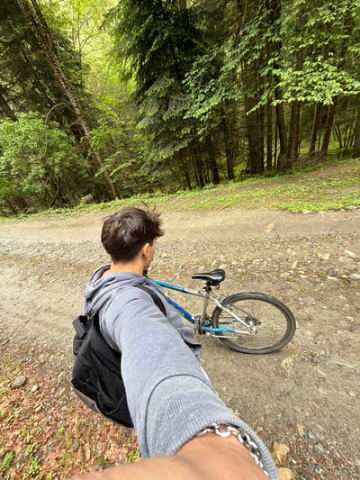 A medium, eye-level shot shows a person with short dark hair and a grey sweatshirt standing on a dirt path next to a bicycle with a blue frame. The person wears a black backpack and a silver bracelet. The bicycle is lying on its side on the path. To the left of the path is a dense forest of tall trees. The path is composed of dirt and small rocks. The location is near Рилски манастир, Bulgaria.