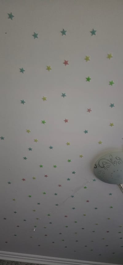 A high-angle, medium shot captures a white ceiling adorned with numerous small, pastel-colored star-shaped stickers. The stars are distributed unevenly across the surface, in shades of light blue, yellow, pink, and green. A light fixture with a patterned glass shade is visible in the lower right quadrant of the image. The ceiling appears to have some minor imperfections or water damage in the central area. The bottom edge of the frame shows the top of a wall with a white decorative molding. The image does not contain any individuals or provide any context related to Karshi City, Uzbekistan.