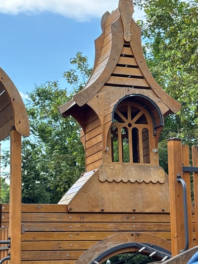 The image shows a close-up of a wooden playground structure in Верхнерусское, Russia. The structure features an ornate wooden tower with a peaked roof and a unique arched window with decorative wooden slats. The tower is built with horizontally placed wooden planks and is supported by vertical wooden posts. In the background, lush green trees are visible against a bright blue sky with scattered white clouds. Part of another wooden playground element is visible on the left side of the frame. A black metal handle and a small heart shape are visible on the right side of the structure. The scene is captured in daylight, suggesting an outdoor setting, likely a park or a dedicated playground area.