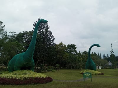 Two large, green dinosaur sculptures, likely made of shrubbery, stand in a grassy park under an overcast sky. The dinosaurs resemble Brachiosaurus or similar long-necked species. Lush trees fill the background, partially obscuring the sky. The foreground includes a sign near the smaller dinosaur. The sculptures are placed on mounds of flowers and greenery, adding to the scene's overall park-like ambiance.