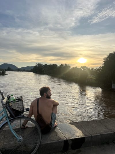 An overhead photograph, taken at sunset, depicts a shirtless male seated on a concrete barrier beside a wide, muddy river in Khorn Neua, Laos. The male, with dark, curly hair and a beard, faces right, looking across the river towards the setting sun. He wears light-colored shorts or swim trunks and a dark brown sling bag across his back. A light blue bicycle with a black wire basket containing two plastic bottles (one white, one green) is positioned to his left, partially visible in the foreground. The river, reflecting golden sunlight, flows from left to right, bordered by dense green foliage on both banks. Multiple tree-covered hills are visible in the distant background under a sky with scattered clouds, displaying warm hues near the sun. The concrete barrier in the immediate foreground appears damp in areas.